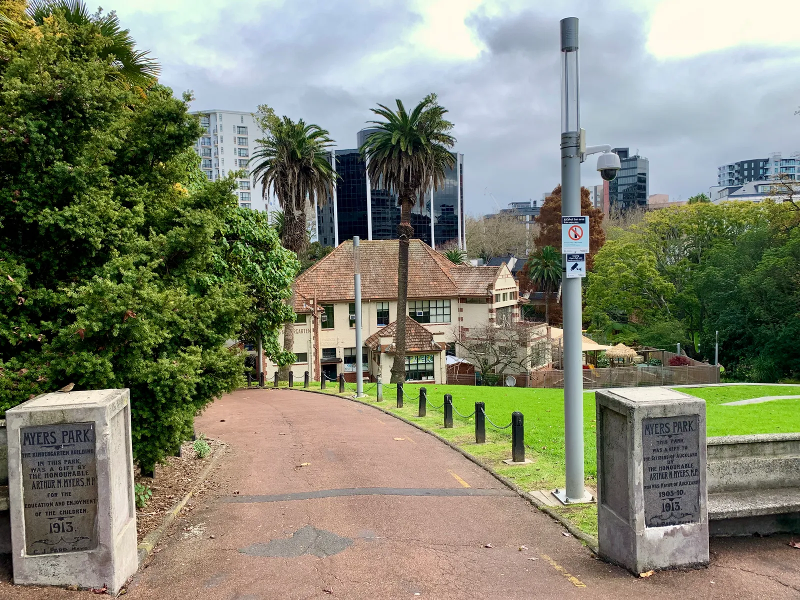 The stone gate piers at the entrance to Myers Park in Auckland, dated 1915, with a path running down between palm trees towards a tiled-roof cottage and city towers behind