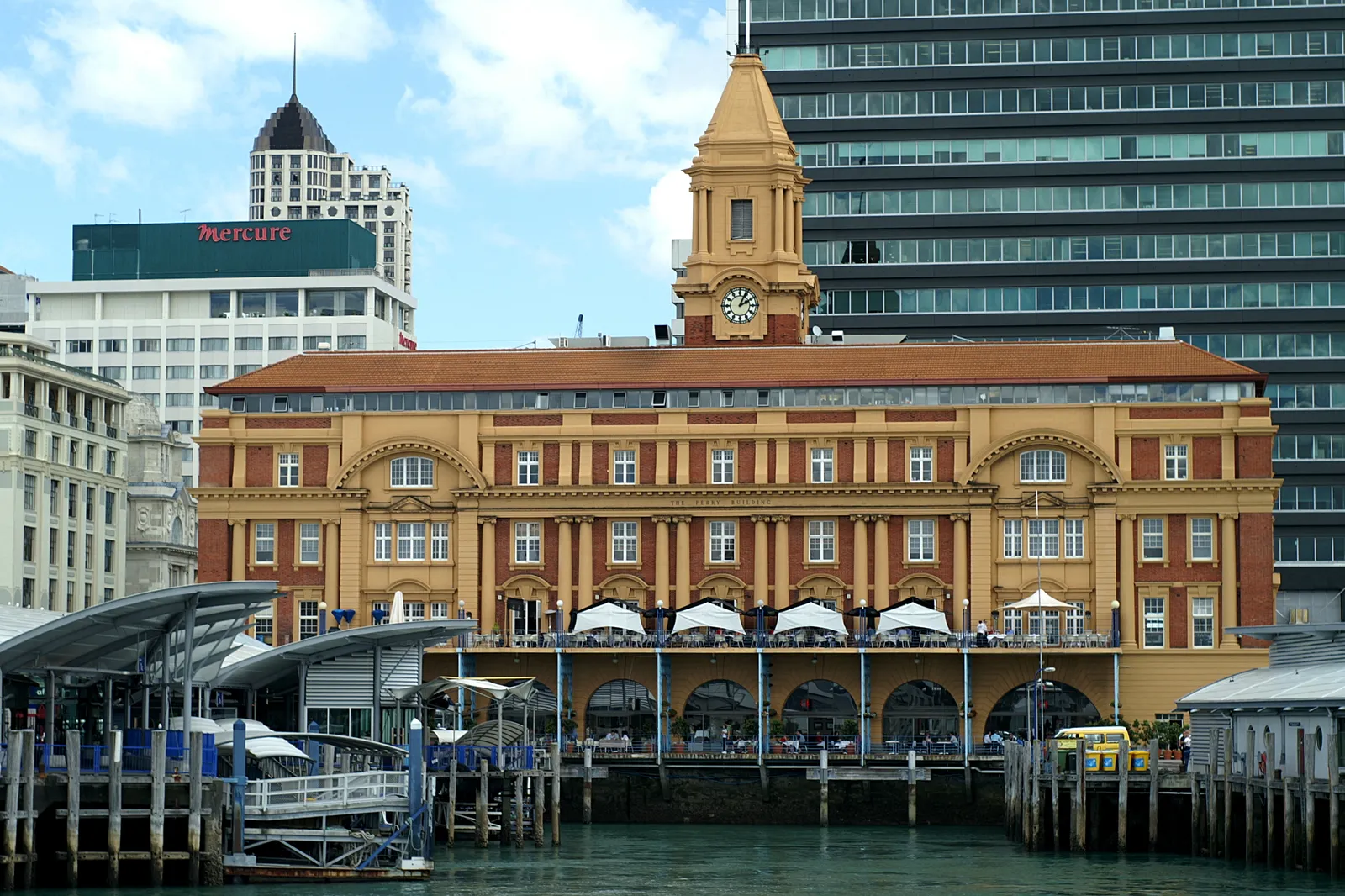 Auckland's Ferry Building seen from the water, a four-storey sandstone block with arched windows and a clock tower, ferry gangways and pontoons along its front