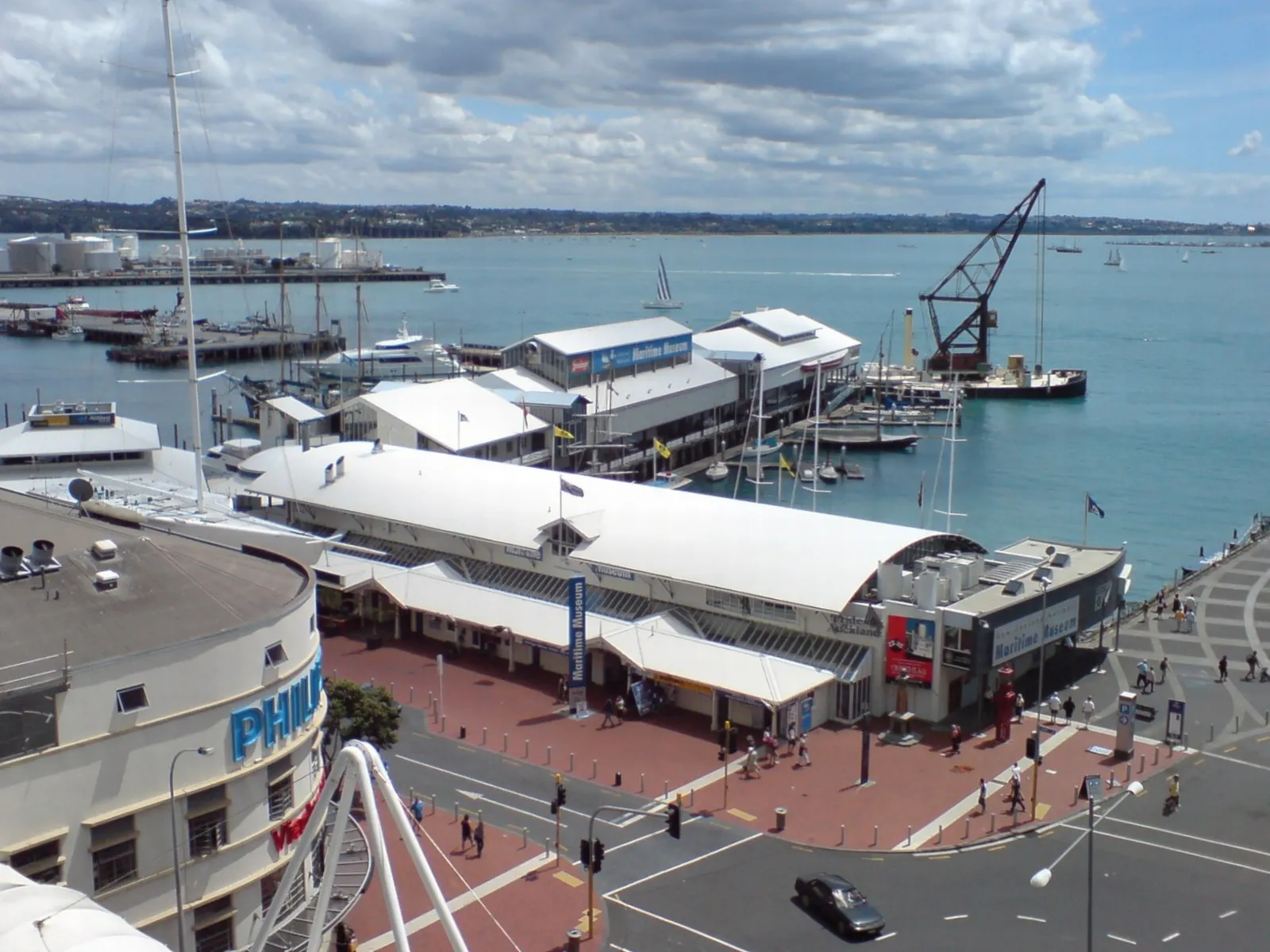 The New Zealand Maritime Museum on Hobson Wharf in Auckland seen from above, its curved white roofs beside the water with masts, a crane and the harbour behind