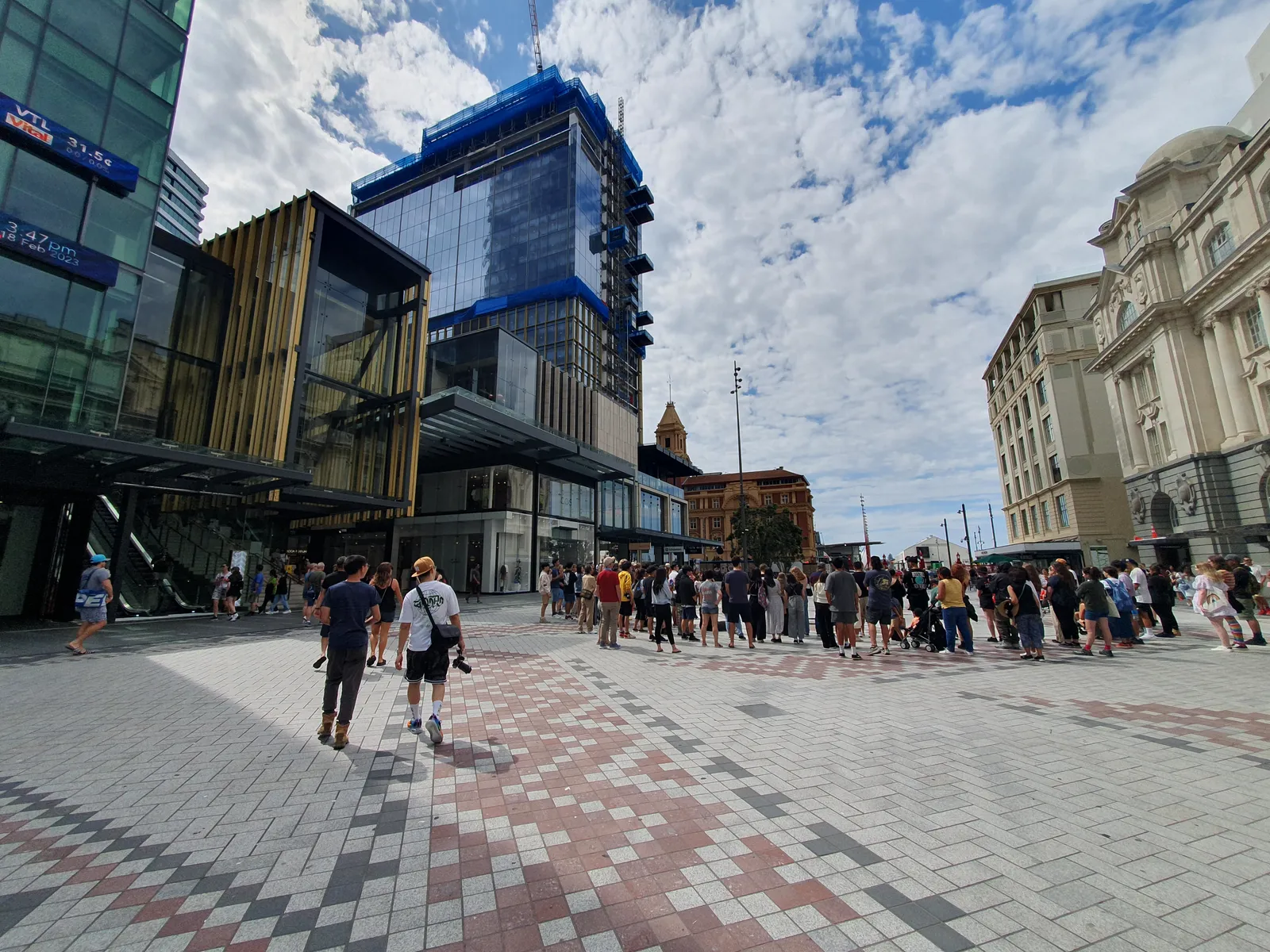 People crossing Te Komititanga in Auckland, a wide public square laid in patterned dark and pale basalt paving, with glass office buildings on one side and older stone facades on the other