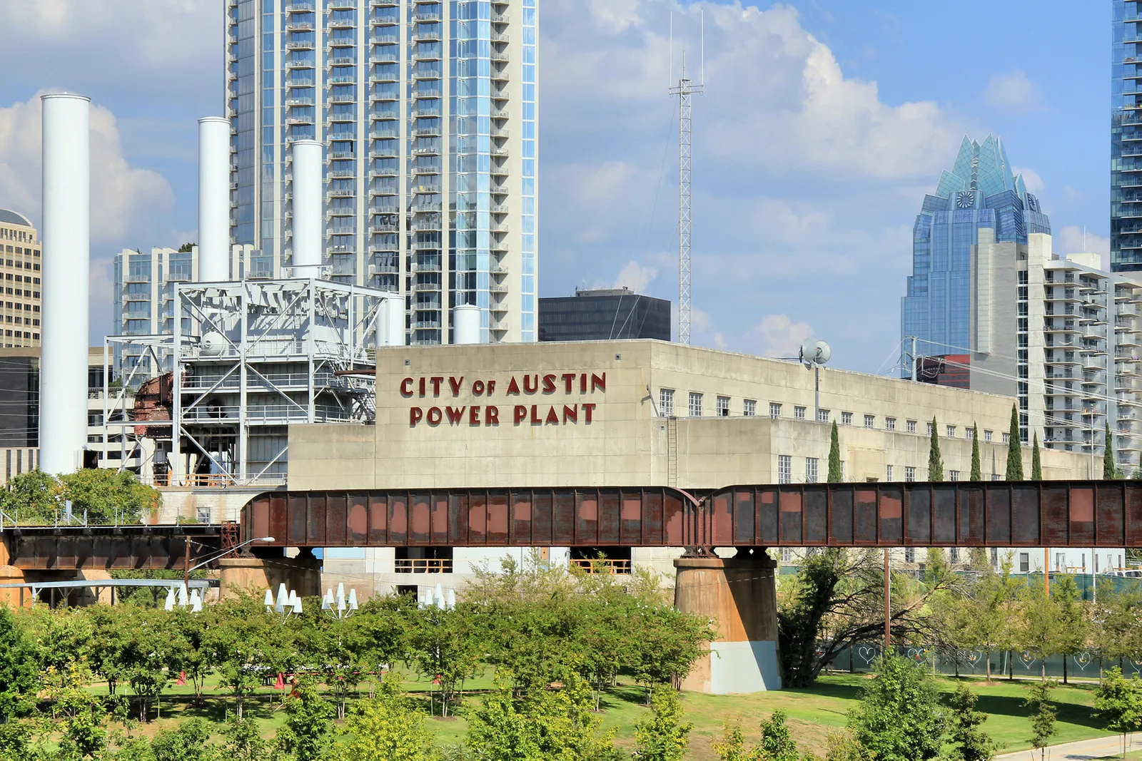 The former Seaholm power plant in Austin, a long pale stone building with CITY OF AUSTIN POWER PLANT lettered across the front, an iron conveyor bridge in front of it and glass towers rising behind
