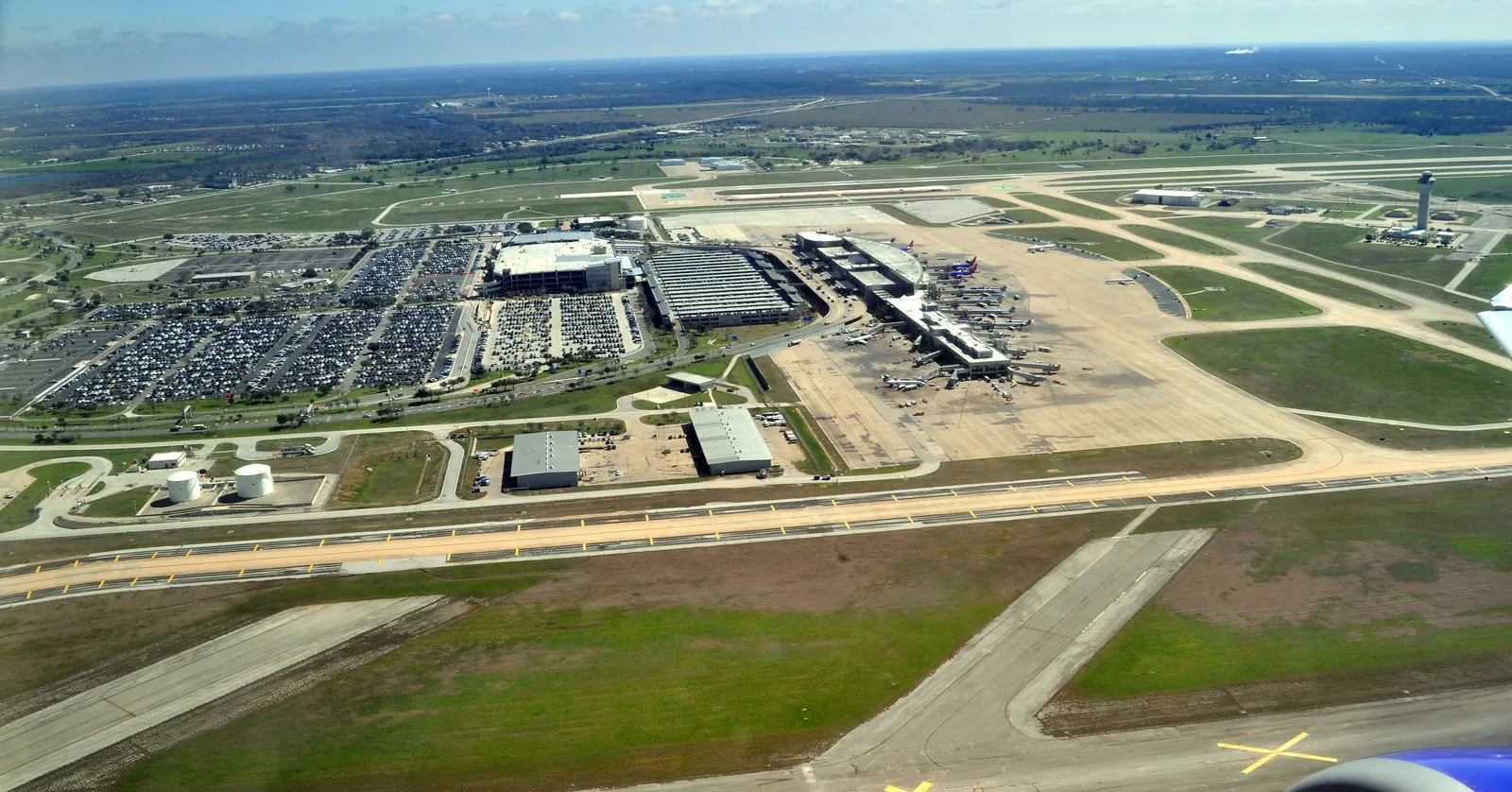 Austin-Bergstrom International Airport from the air, a long low terminal with jets parked nose-in along its pier, runways and taxiways stretching across flat green land to the horizon