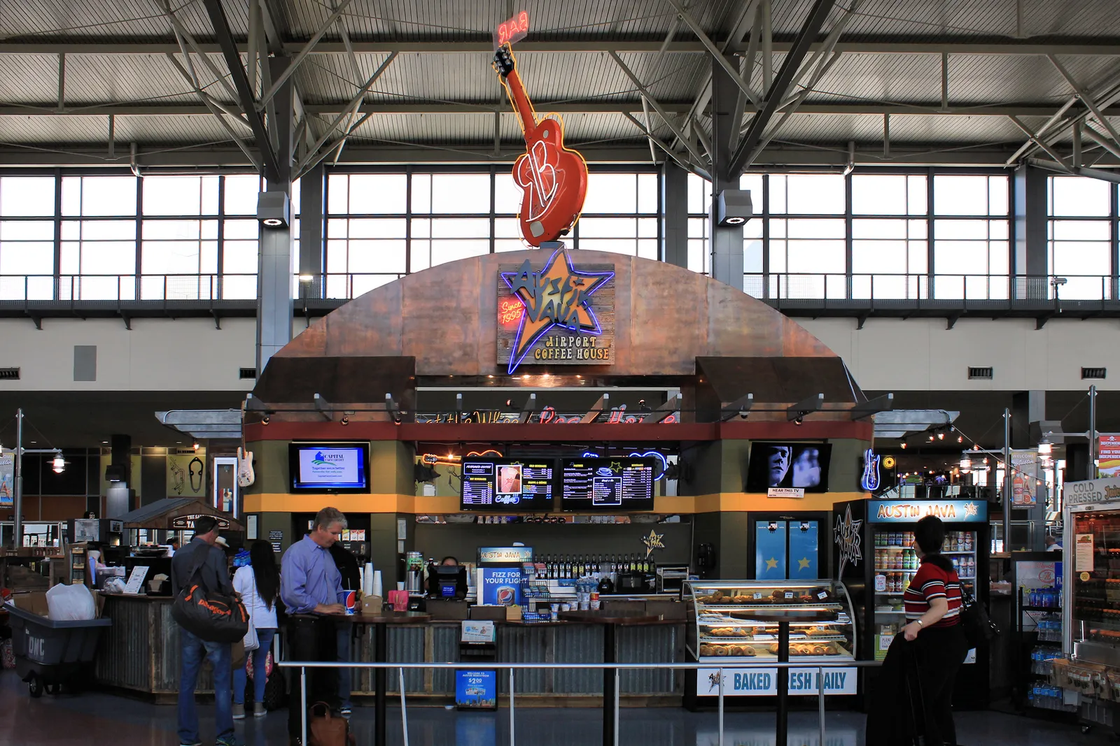 A food and coffee stand on the concourse at Austin's airport, a curved copper-clad counter under a neon guitar and a lit Texas star sign, with customers waiting at the till