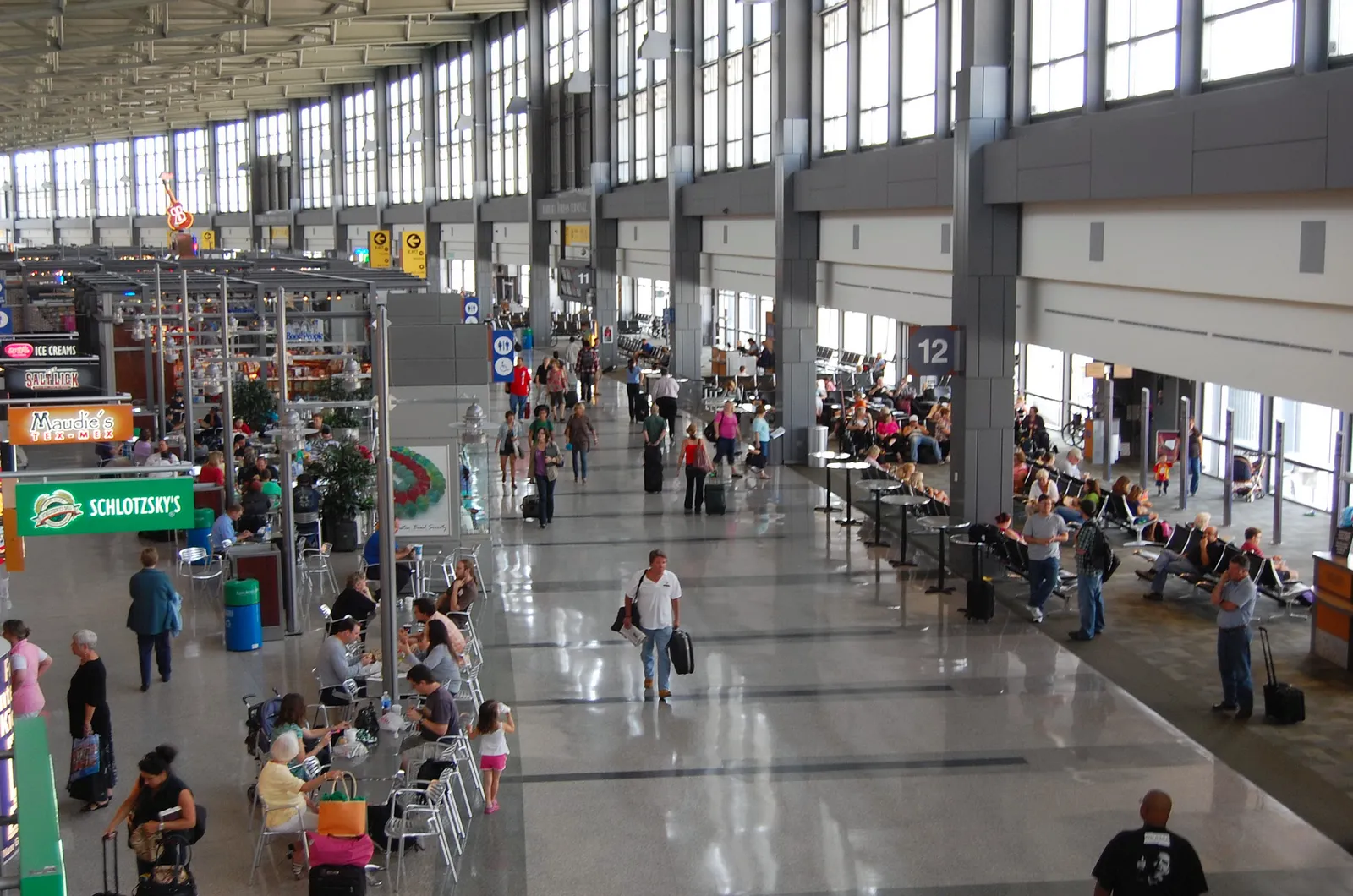 The concourse inside Austin's Barbara Jordan Terminal seen from above, a long hall with a glass wall of windows down one side, gate seating on the right and a row of food and drink stands on the left, passengers walking through the middle