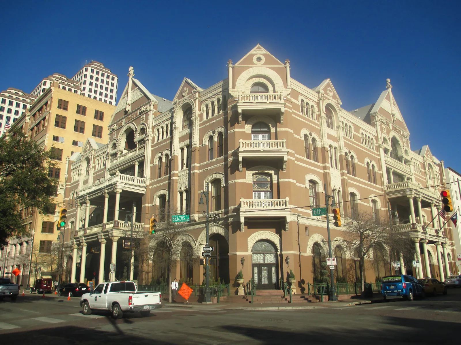 The Driskill Hotel on the corner of Sixth and Brazos in Austin, a four-story brick and limestone building with arched windows, balconies and steep gables, seen from the street crossing