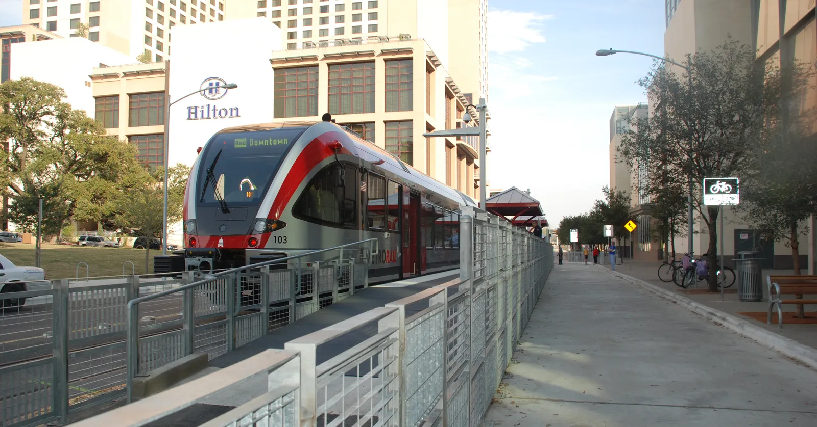 A red and grey commuter train standing at the open-air Downtown Station platform in Austin, with a metal railing along the sidewalk beside it and downtown buildings rising behind