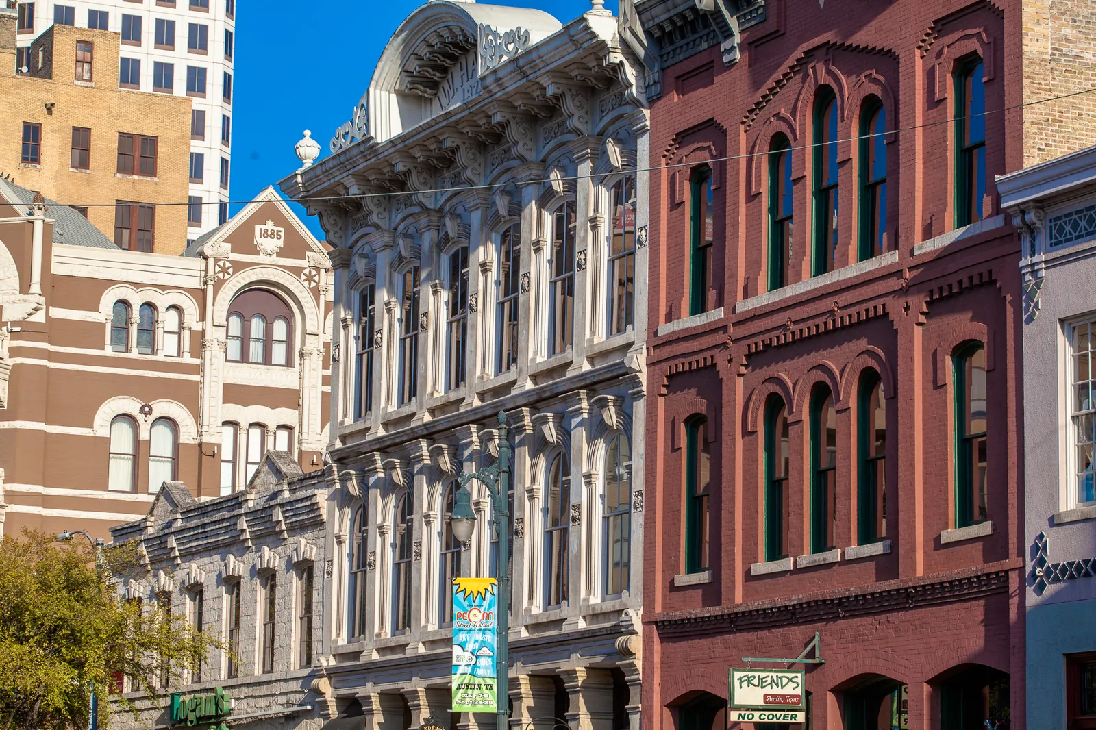 A row of 19th-century commercial buildings on Sixth Street in Austin, brick and painted stone fronts with arched windows, decorated cornices and iron balconies, a glass tower rising behind them