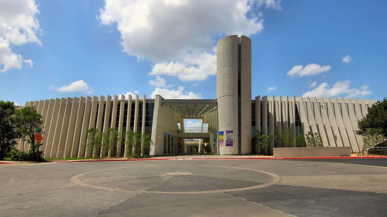 The Emma S. Barrientos Mexican American Cultural Center in Austin, a long white concrete building of vertical fins with a round drum tower at its center and a paved forecourt in front