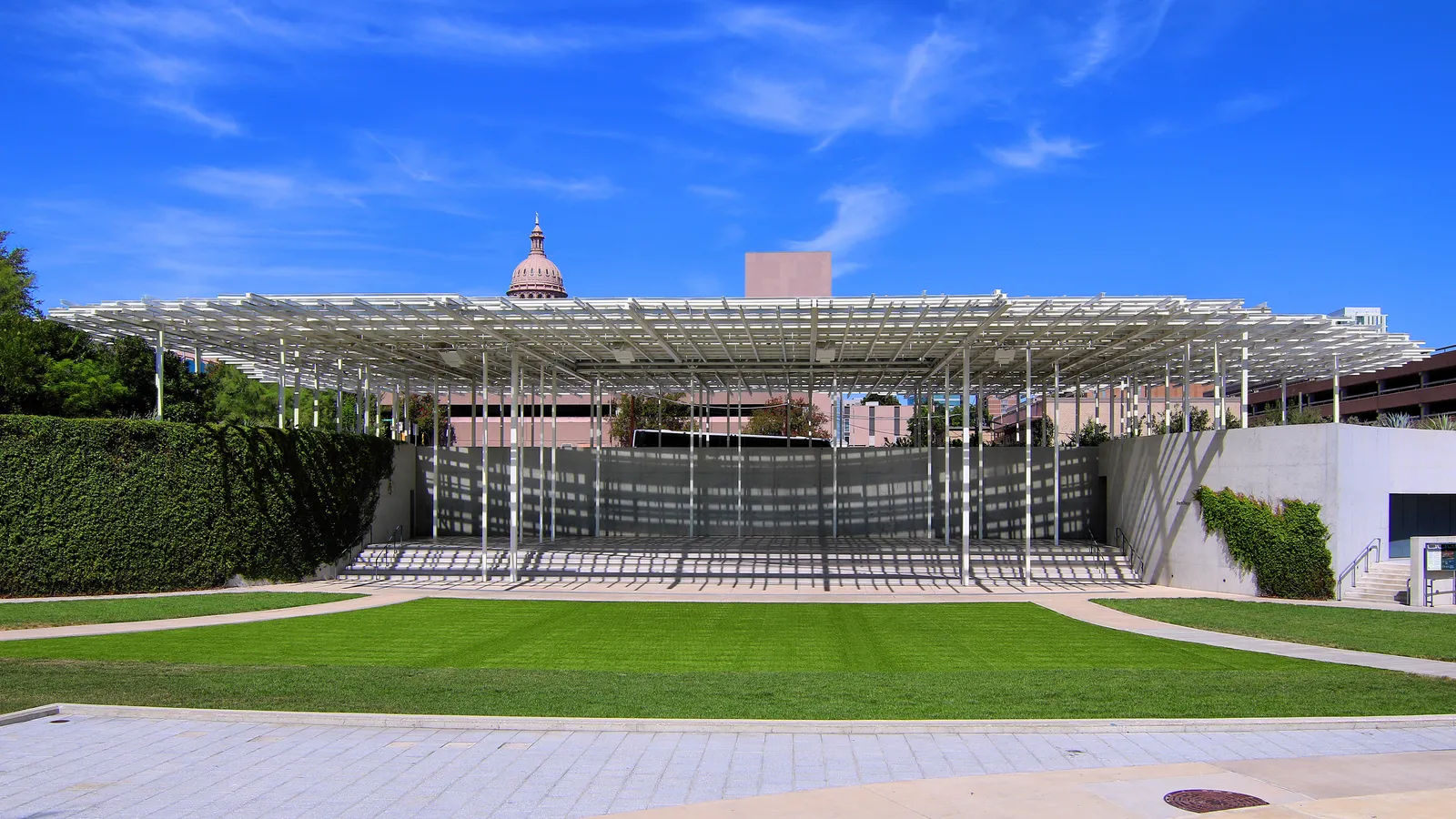 The Moody Amphitheater in Waterloo Park in Austin, a wide white lattice canopy on slim columns over banked seating and an open lawn, with the Capitol dome visible behind it