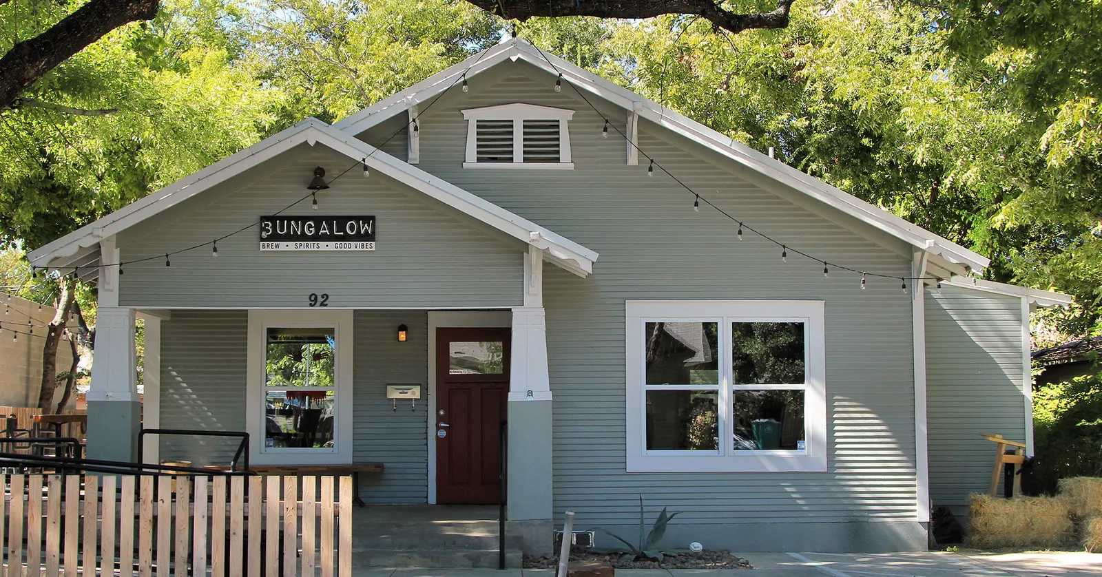 A pale green wooden bungalow on Rainey Street in Austin turned into a bar, with a gabled porch, a hand-painted sign over the steps, string lights across the eaves and a picket fence in front
