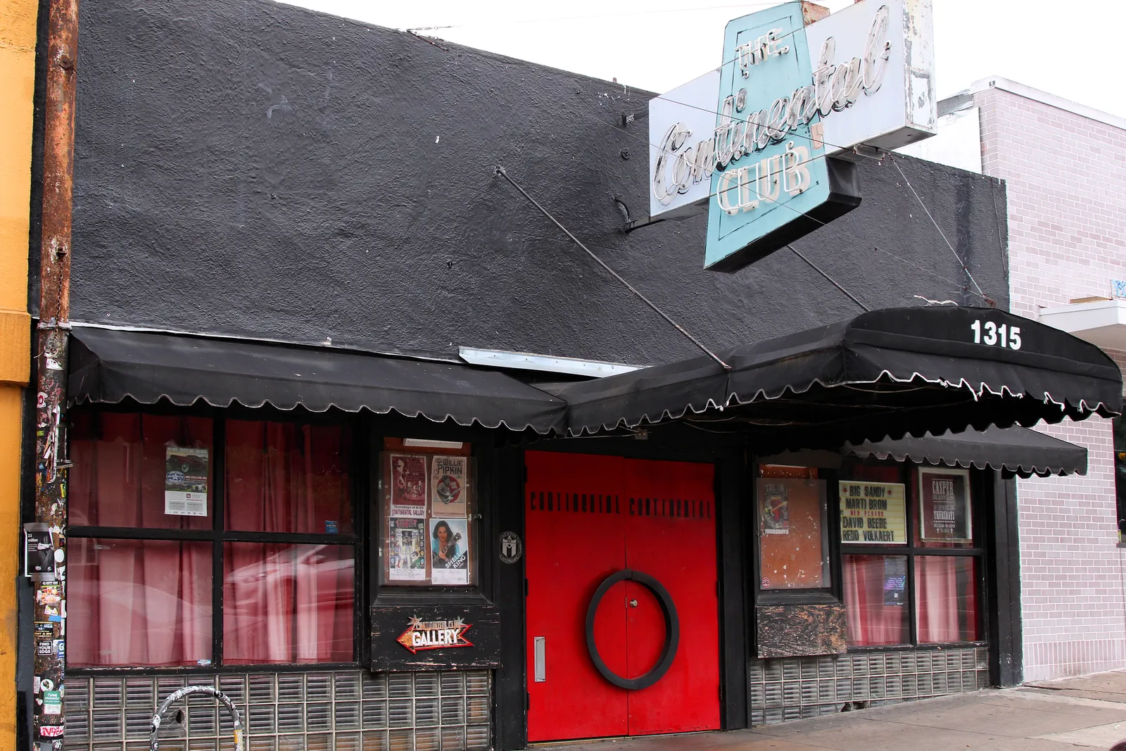 The front of the Continental Club at 1315 South Congress in Austin, a low black-painted single-story building with a red door, red curtained windows, scalloped awnings and a vertical neon sign above