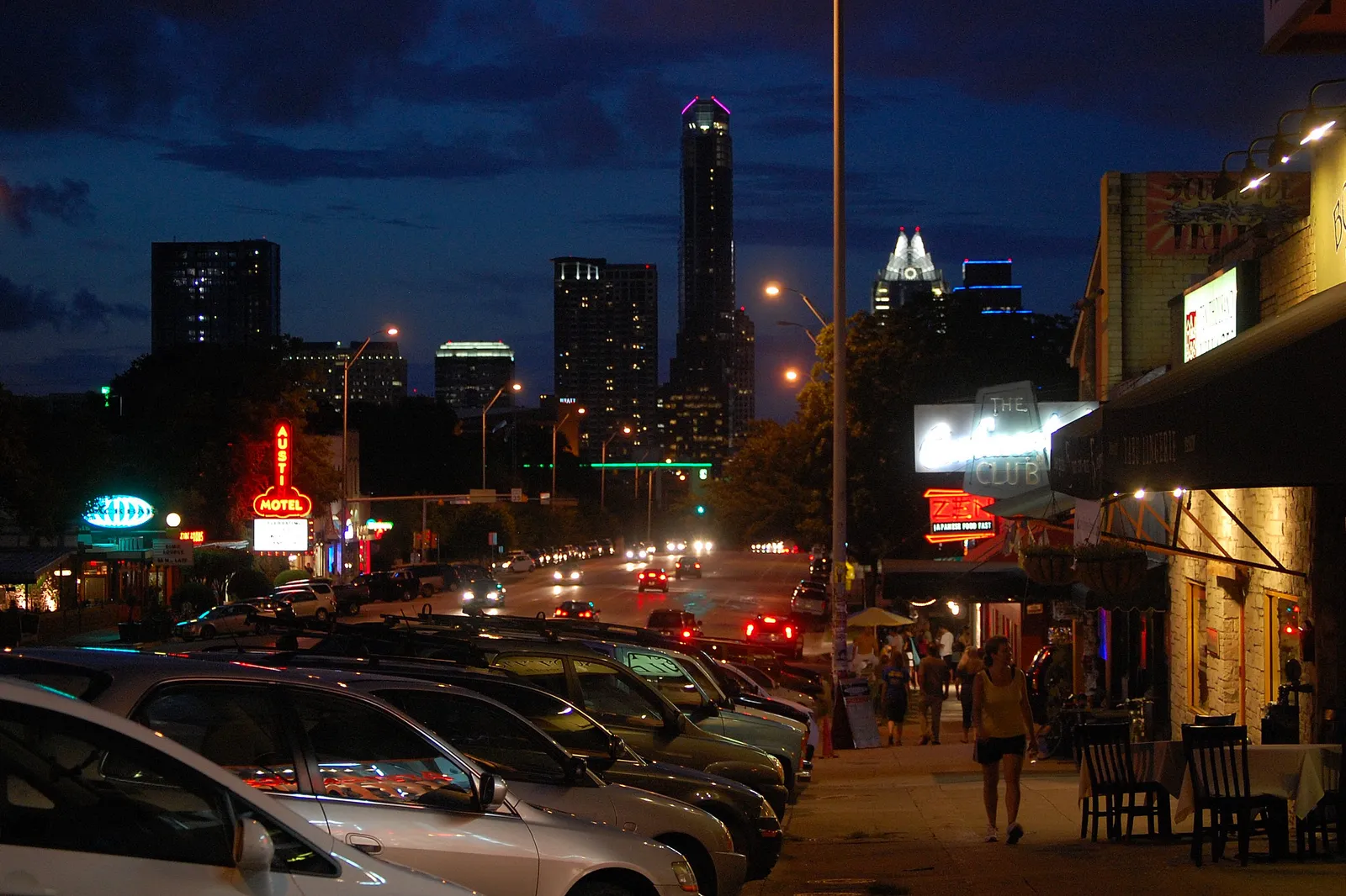 South Congress Avenue in Austin at dusk, neon motel and shop signs lit along both sides of the street, parked cars in the foreground and the downtown skyline and lit Capitol dome on the horizon
