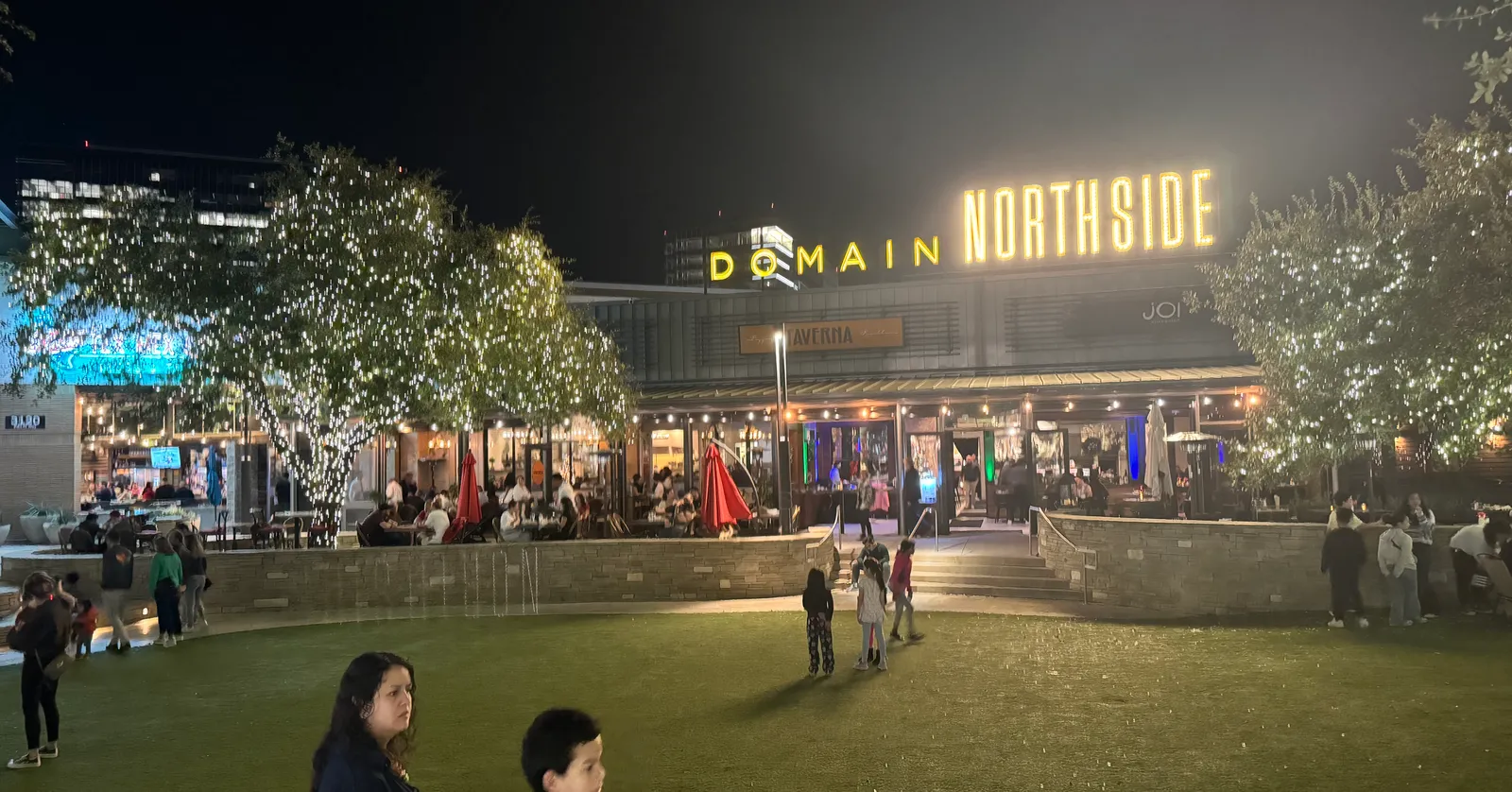 Domain Northside in Austin after dark, a lit sign reading DOMAIN NORTHSIDE above a low block of restaurants, with string lights through the trees and people crossing a lawn in front