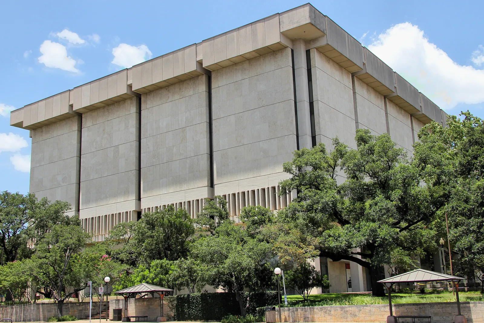 The Harry Ransom Center in Austin, a large windowless concrete block set on a colonnaded base with narrow vertical fins, trees and a small pavilion in front of it