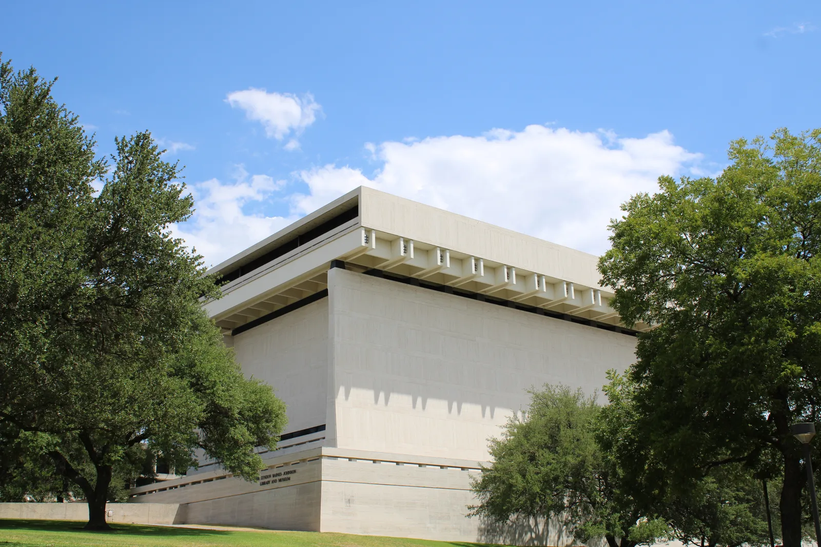 The Lyndon B. Johnson Presidential Library in Austin, a windowless travertine box with a deep overhanging roof standing on a grass rise between live oaks under a blue sky