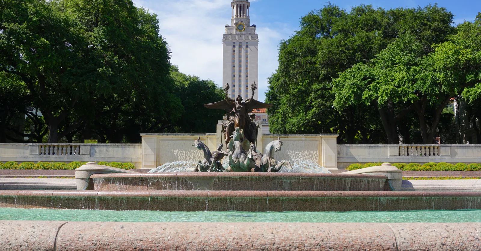 The University of Texas Tower in Austin rising pale above a line of live oaks, with the bronze figures and basin of the Littlefield Fountain in the foreground and water running over its steps