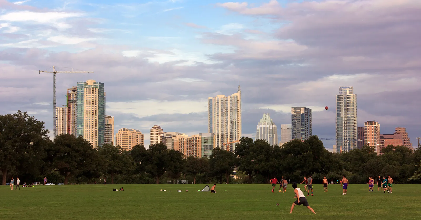 The open Great Lawn at Zilker Park in Austin on a summer evening, people playing on the mown grass in the foreground and the downtown skyline standing above a line of trees behind