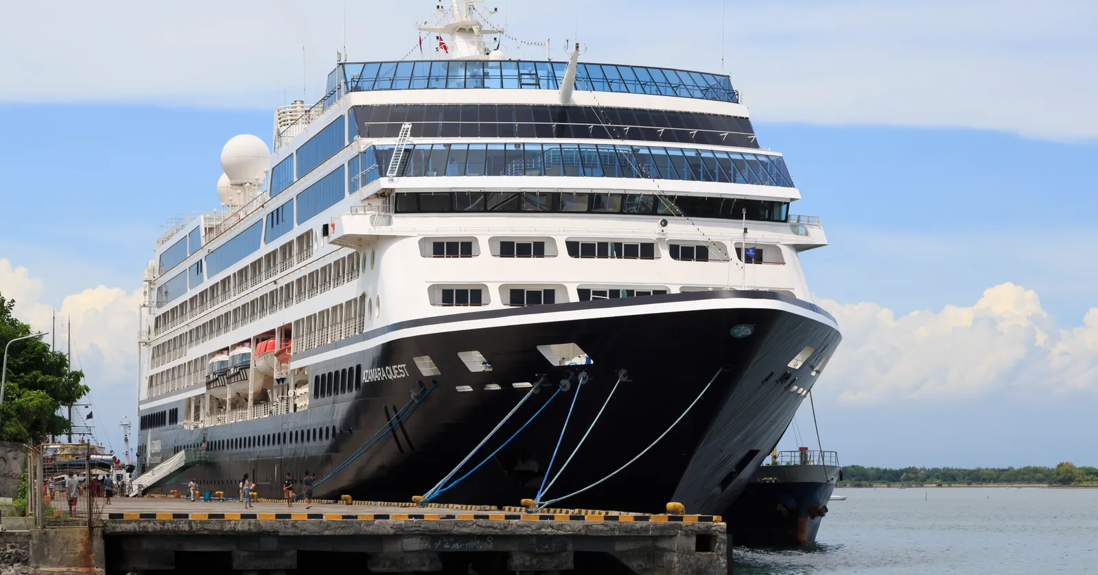 A white cruise ship berthed bow-on against the concrete quay at Benoa in Bali, with a gangway down to the pier and flat water beyond