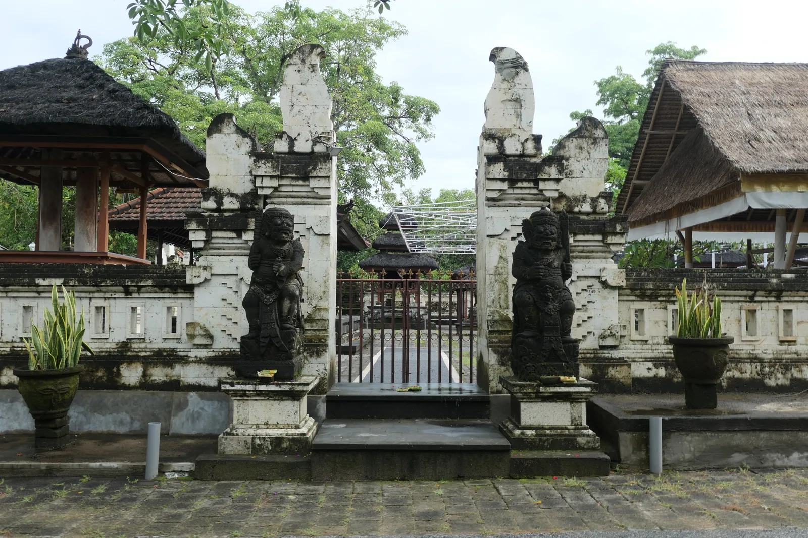 The split stone gateway of Pura Sakenan on Serangan in Bali, weathered pale, with carved guardian figures either side and thatched pavilions behind the wall