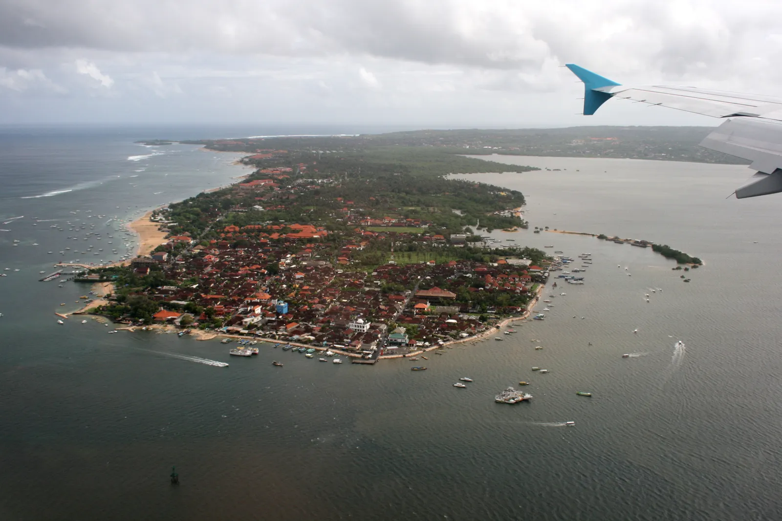 The Tanjung Benoa peninsula in Bali seen from a departing aircraft, a dense red-roofed strip of land narrowing into the bay with boats moored along both shores