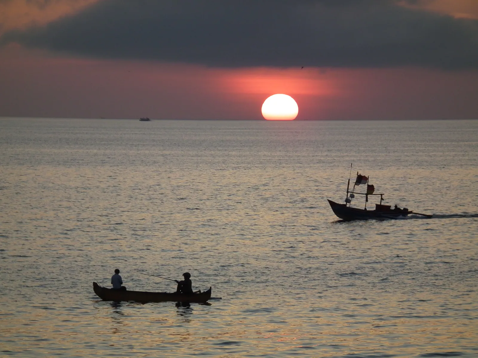 The sun dropping into the sea off Jimbaran Bay in Bali, silhouetting a small outrigger with two people in it and a motorised fishing boat further out