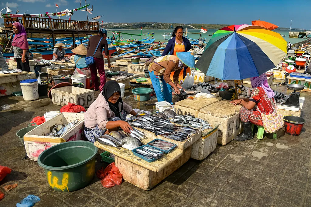 Sellers laying out fish on concrete slabs under a striped umbrella at the Kedonganan fish market in Bali, with moored outrigger boats and the bay behind them
