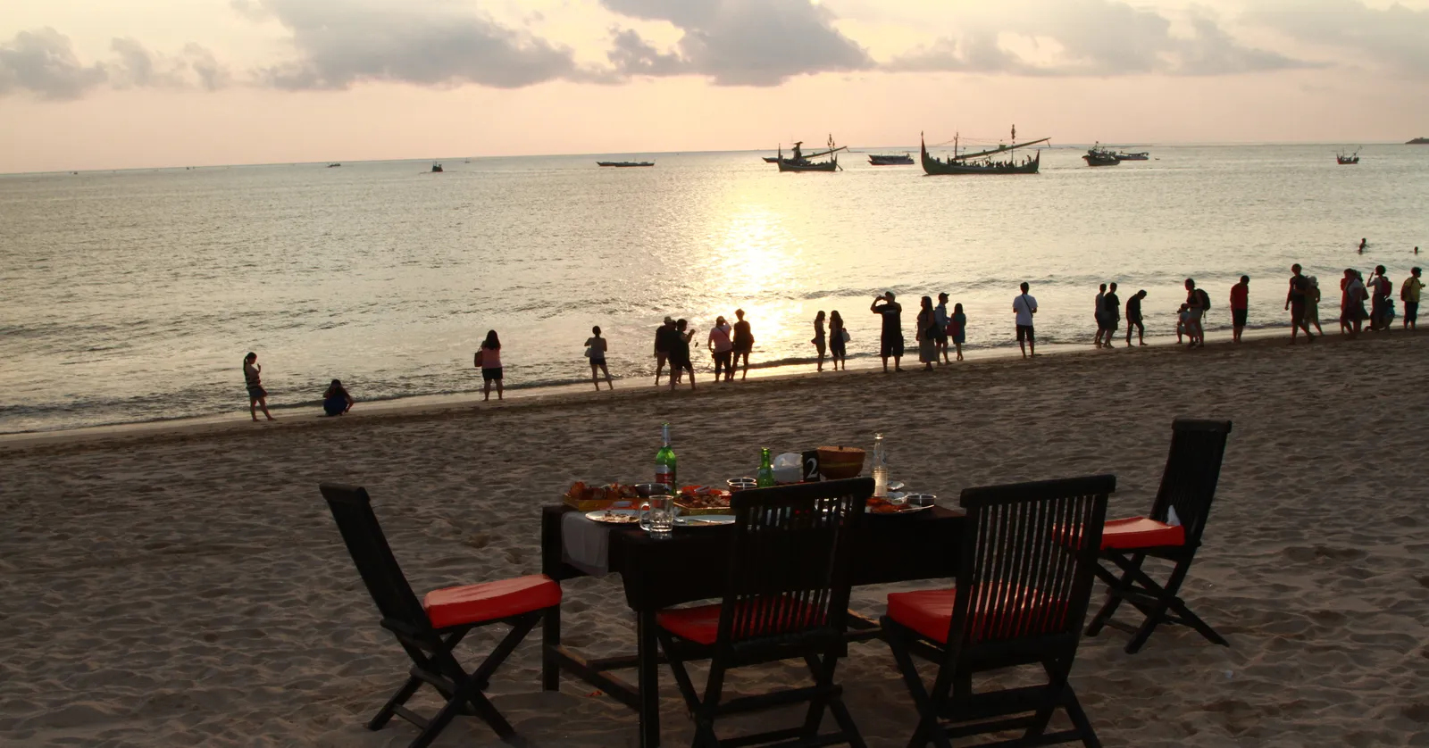 A laid dinner table with red-cushioned wooden chairs standing directly on the sand at Jimbaran Bay in Bali, with people along the waterline and fishing boats offshore as the sun goes down