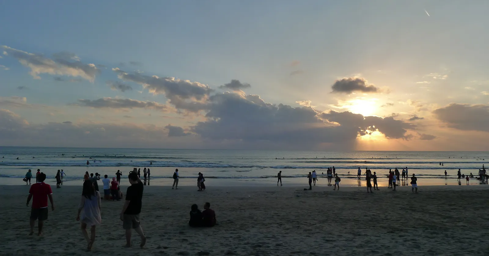 Crowds standing and sitting on the wide flat sand of Kuta Beach in Bali as the sun drops behind broken cloud over the surf