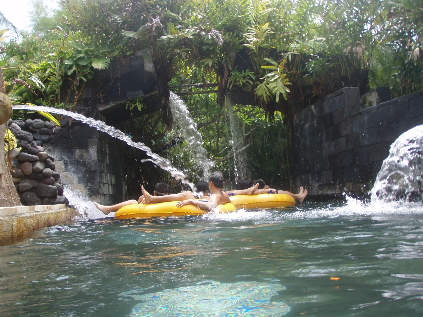 Two people floating on yellow rings down the lazy river at the Kuta water park, past stone walls with waterfalls pouring over them under dense tropical planting