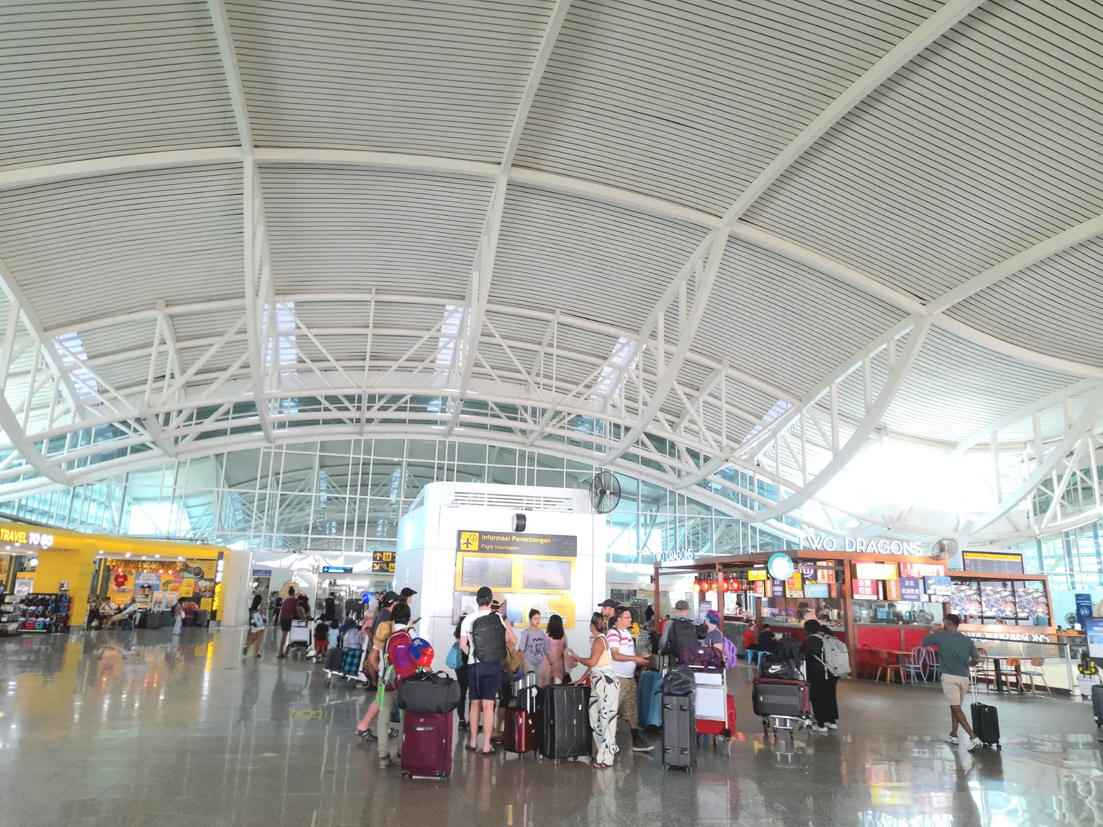 The departure hall of the international terminal at Bali's airport, with a curved white ribbed roof on branching steel columns above passengers queueing with trolleys