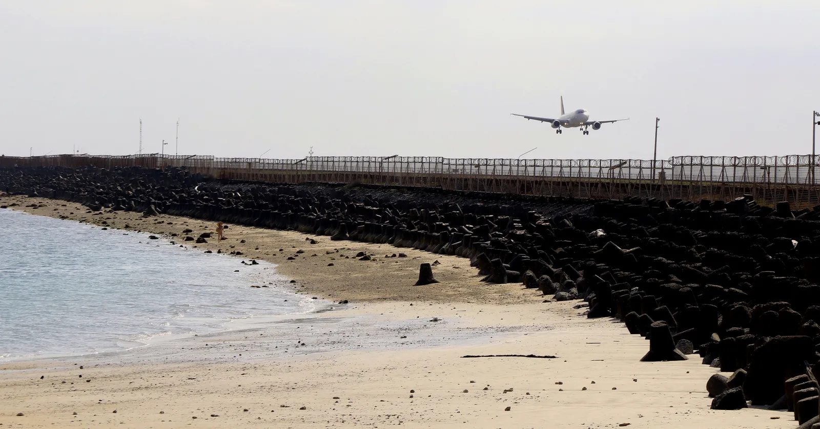 An airliner on final approach over the black rock seawall at the end of the runway at Bali's airport, with the empty sand of Kelan Beach in the foreground