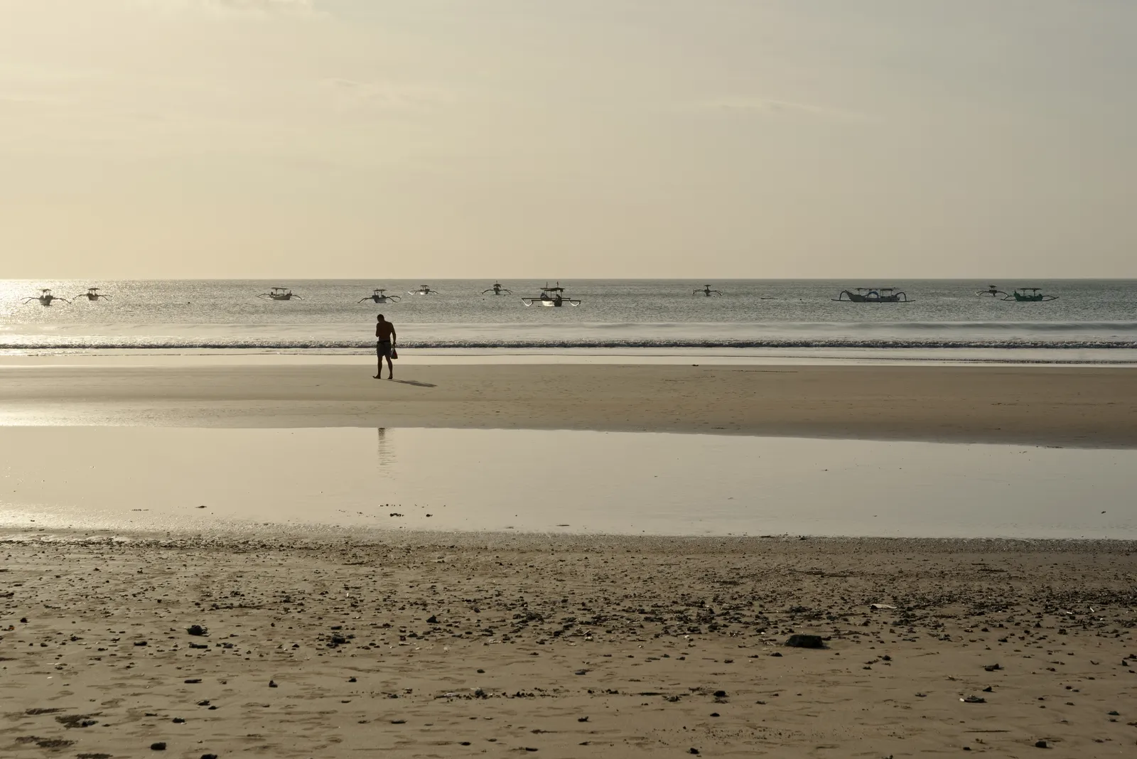 Wide wet sand at the southern end of Kuta Beach in Bali reflecting the low sun, with a single figure walking and outrigger fishing boats moored offshore
