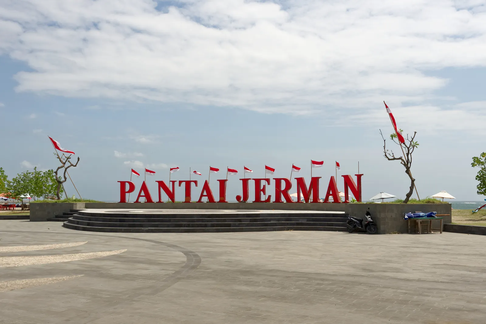 Large red letters spelling PANTAI JERMAN on a paved forecourt above the beach in Tuban, Bali, with a row of Indonesian flags behind them and the sea beyond