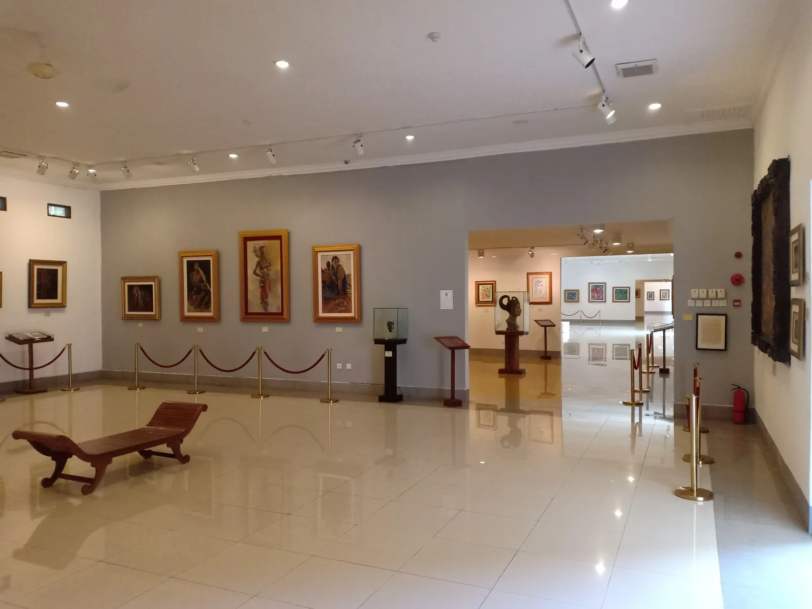 A gallery at Museum Pasifika in Nusa Dua, framed paintings hung along grey and white walls above a polished floor with rope barriers and a carved bench