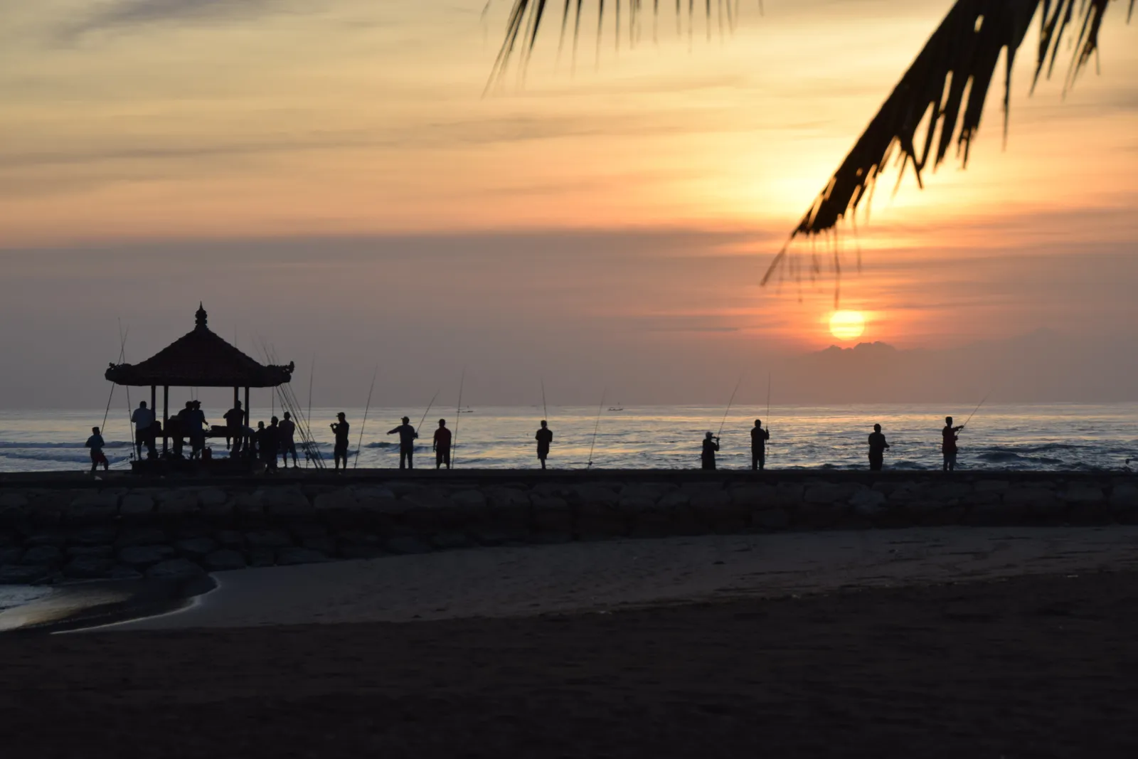 A thatched bale and a line of people fishing from a low stone jetty at Nusa Dua beach in Bali, silhouetted against the sun low over flat water
