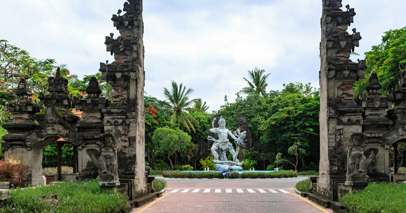 The split candi bentar gate into Nusa Dua in Bali, two carved stone halves either side of the road with a stone fountain statue on the roundabout behind them