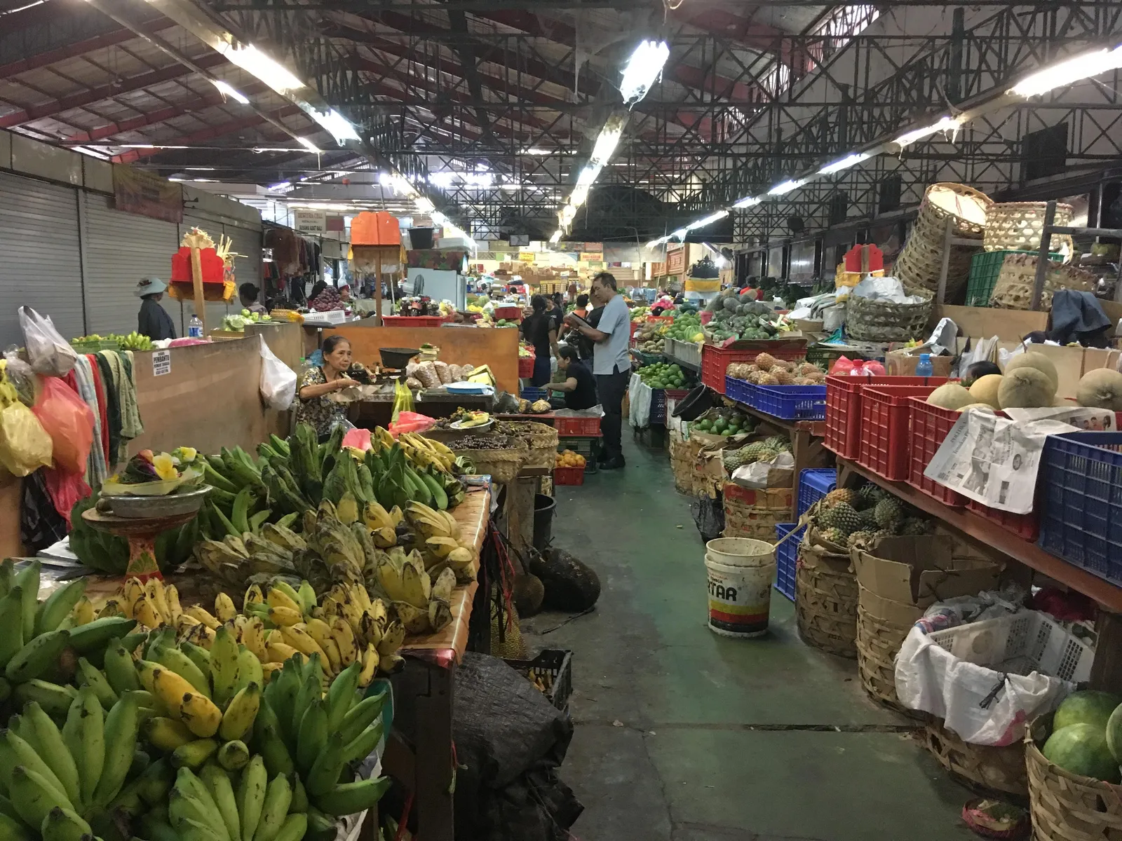 An aisle inside Pasar Badung in Denpasar lined with stacked bananas, papayas and vegetables on wooden tables, with traders working under strip lighting