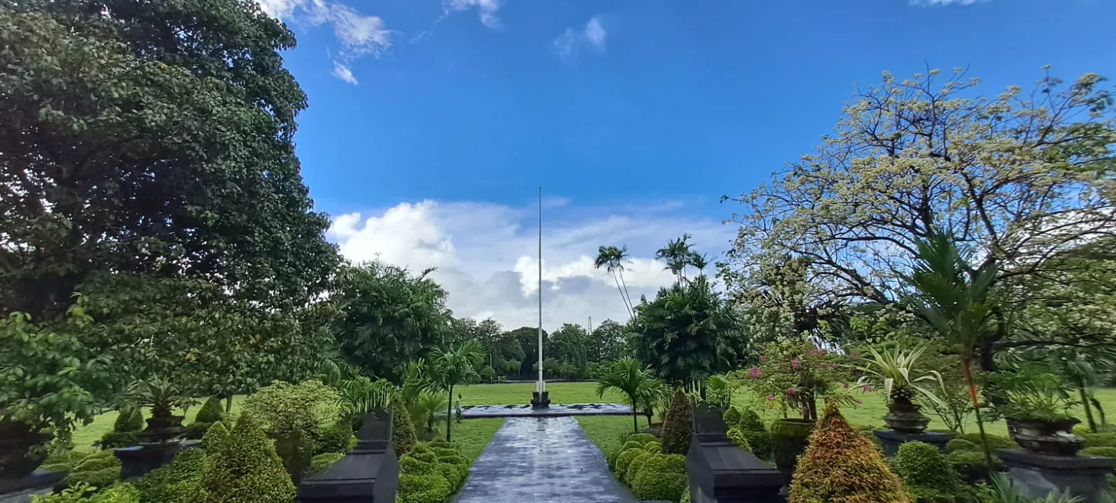 The green lawns, clipped shrubs and central flagpole of Puputan Badung Square in Denpasar, with big shade trees on both sides under a bright sky