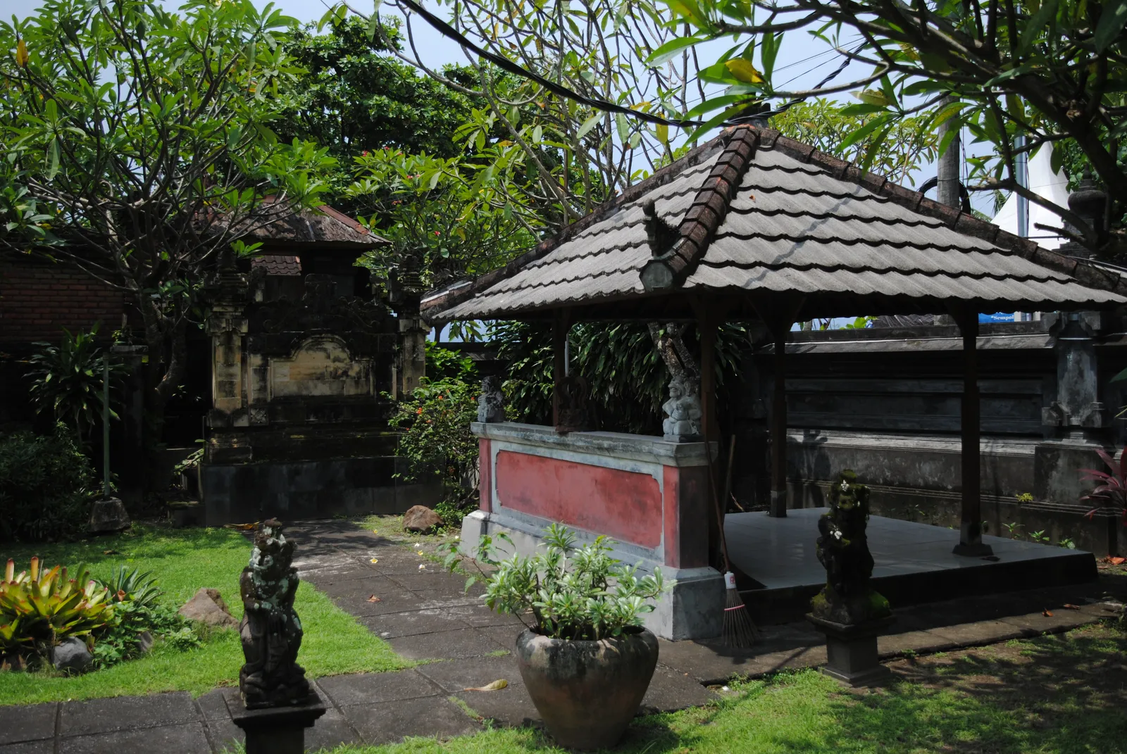 A tiled Balinese pavilion with a red-panelled platform standing in the planted garden of the Le Mayeur house museum in Sanur, with carved guardian figures on the lawn