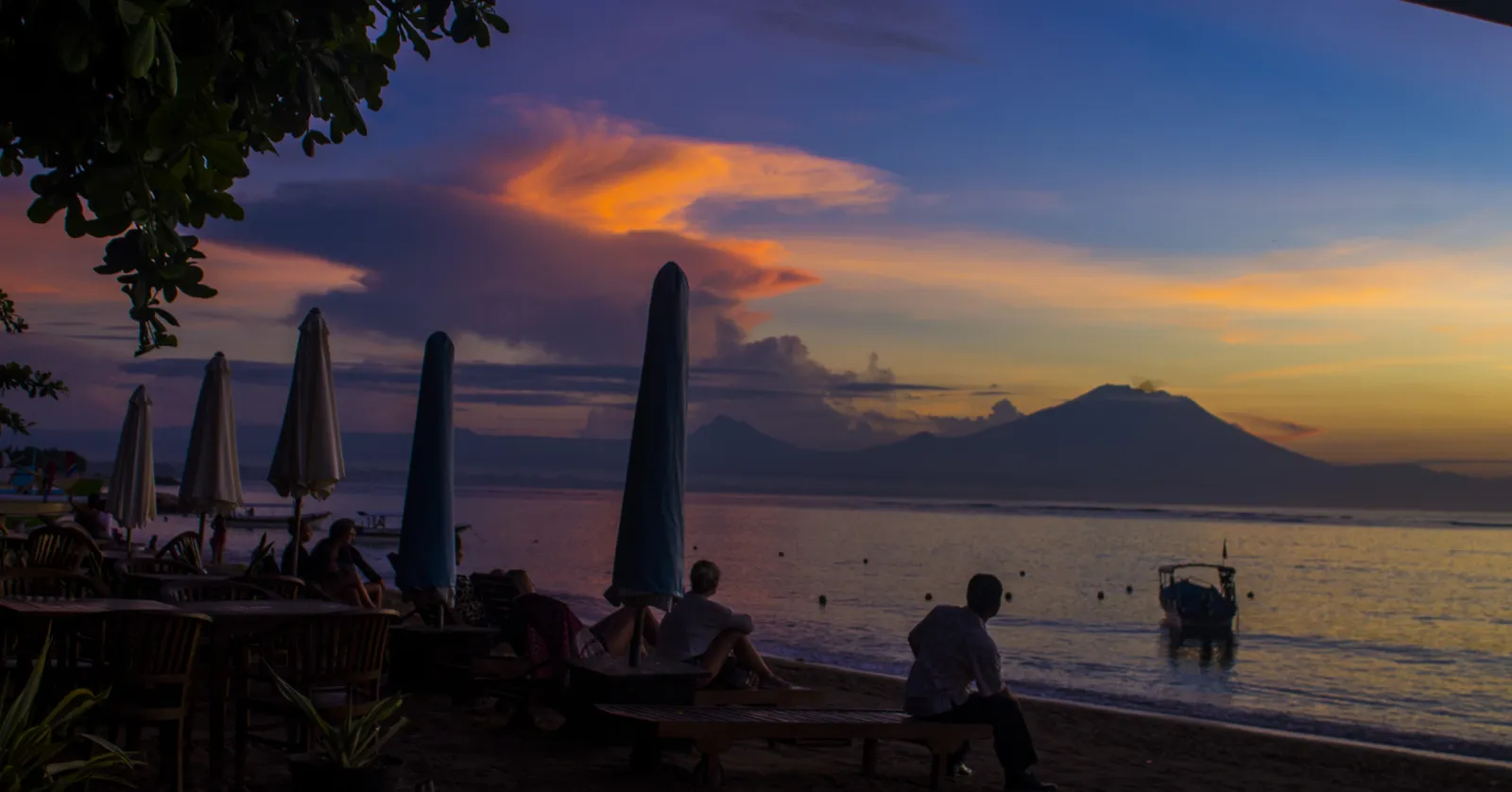 Furled umbrellas and people sitting on benches along the sand at Sanur in Bali, with colour in the sky over flat water and a volcano silhouetted on the far shore