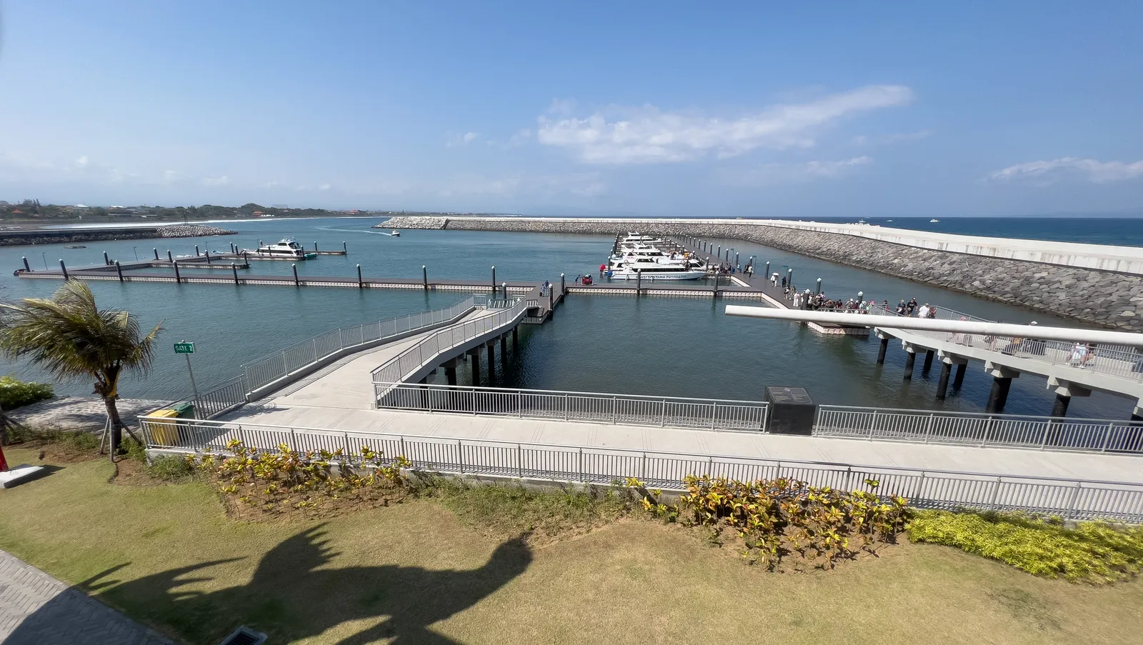 The concrete breakwater and floating pontoons of Sanur harbour in Bali, with fast boats moored along the jetties and passengers walking the walkway ashore