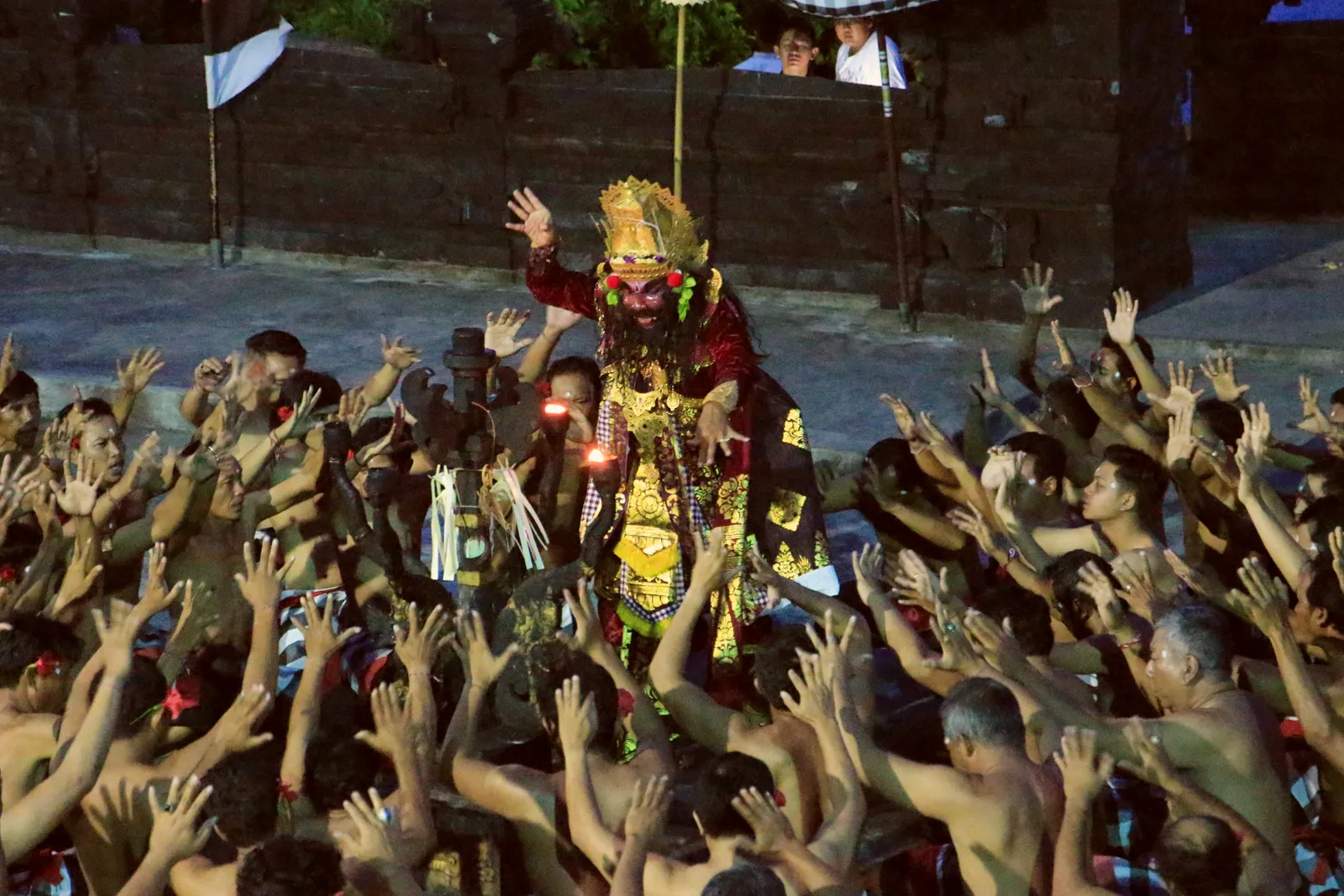 A masked and crowned performer standing with raised hands at the centre of the Kecak circle at Uluwatu, ringed by dozens of seated chanters with their arms in the air