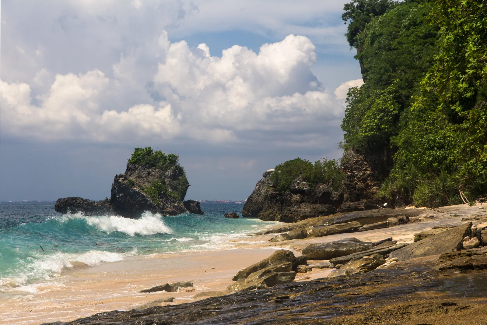 The small sandy cove of Padang Padang Beach in Bali, hemmed in by overhanging limestone and jungle with a rock stack standing in the surf offshore