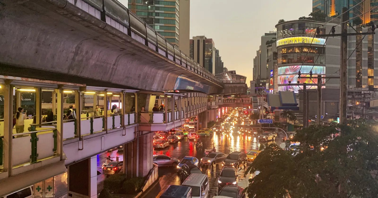 Sukhumvit Road at Asok in Bangkok at dusk, the elevated railway running along the left above a covered walkway with lit shopfronts and standing traffic filling the road below