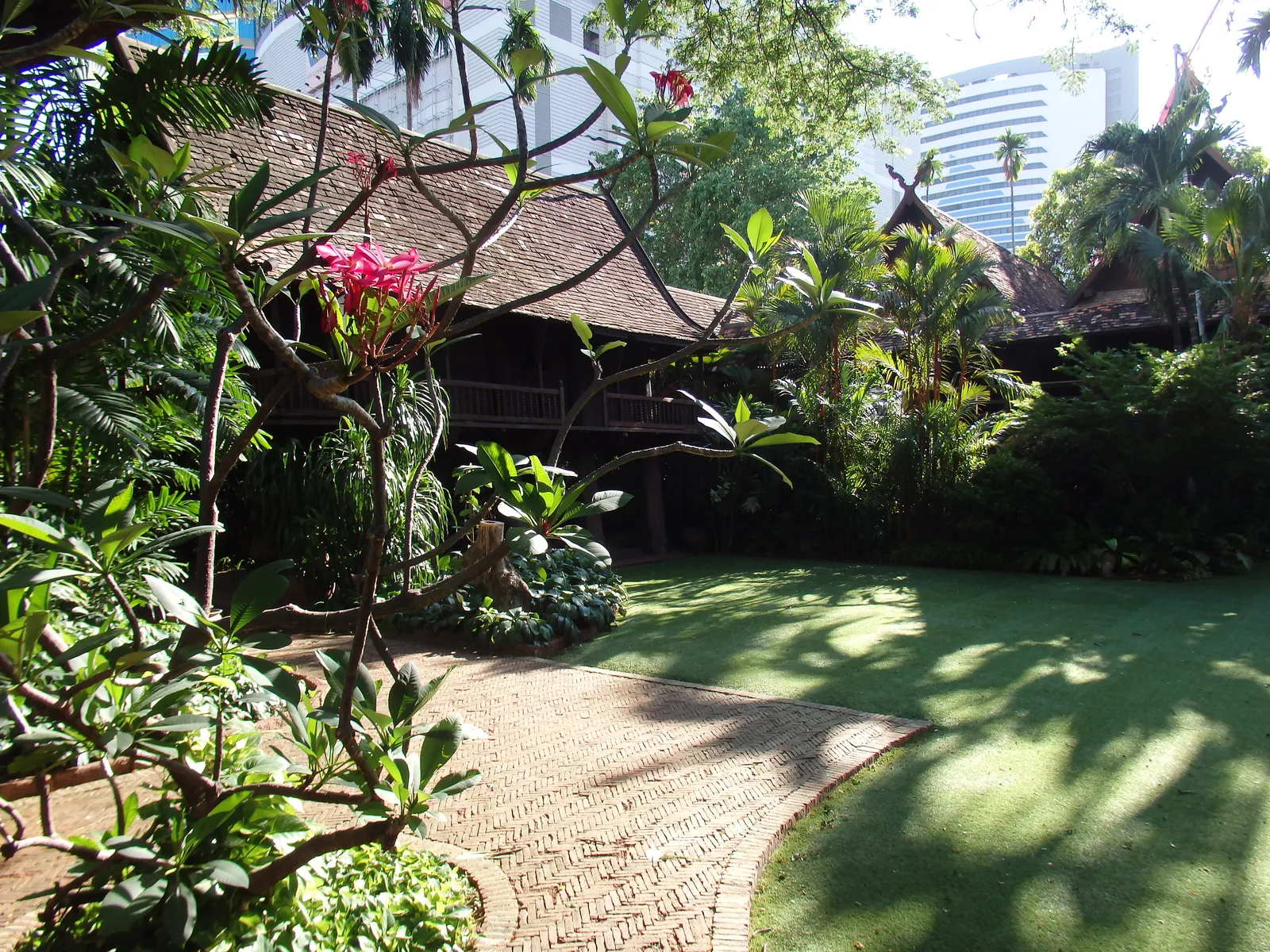 Kamthieng House in Bangkok, a dark teak house on posts under a steep tiled roof at the end of a lawn, screened by frangipani and palms with a modern office tower rising behind it
