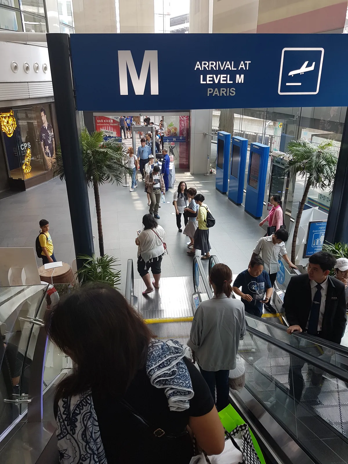 Inside Terminal 21 in Bangkok, an airport-style blue sign reading arrival at level M, Paris, hung over escalators carrying shoppers down between palm-planted floors