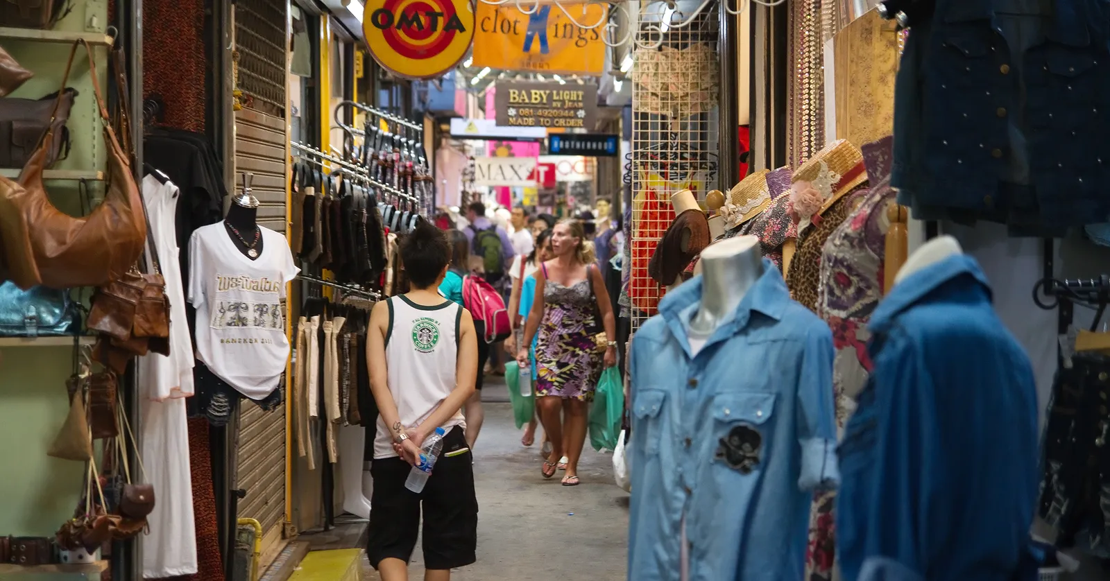 A narrow alley inside Chatuchak Weekend Market in Bangkok, clothing and bags hung from both sides of a covered walkway barely wide enough for two people to pass