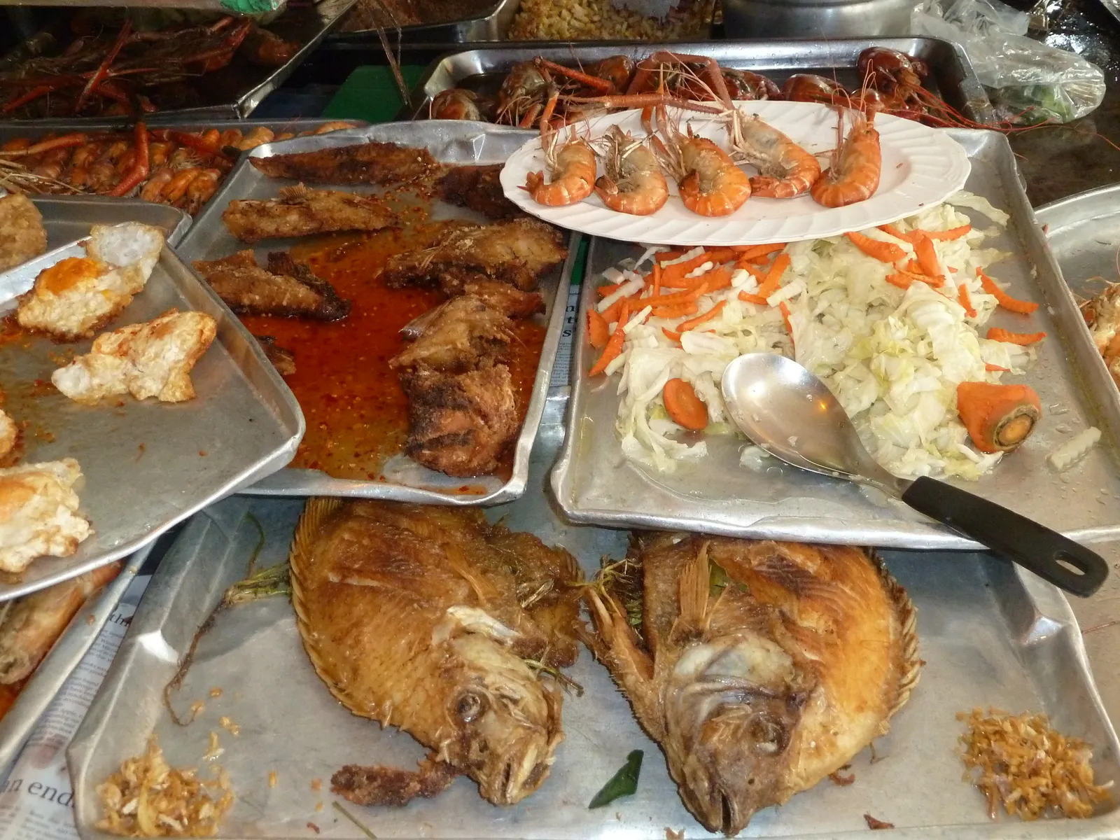 A food stall counter at Chatuchak Weekend Market in Bangkok, steel trays of whole fried fish, grilled prawns and shredded papaya salad laid out with a serving spoon across them