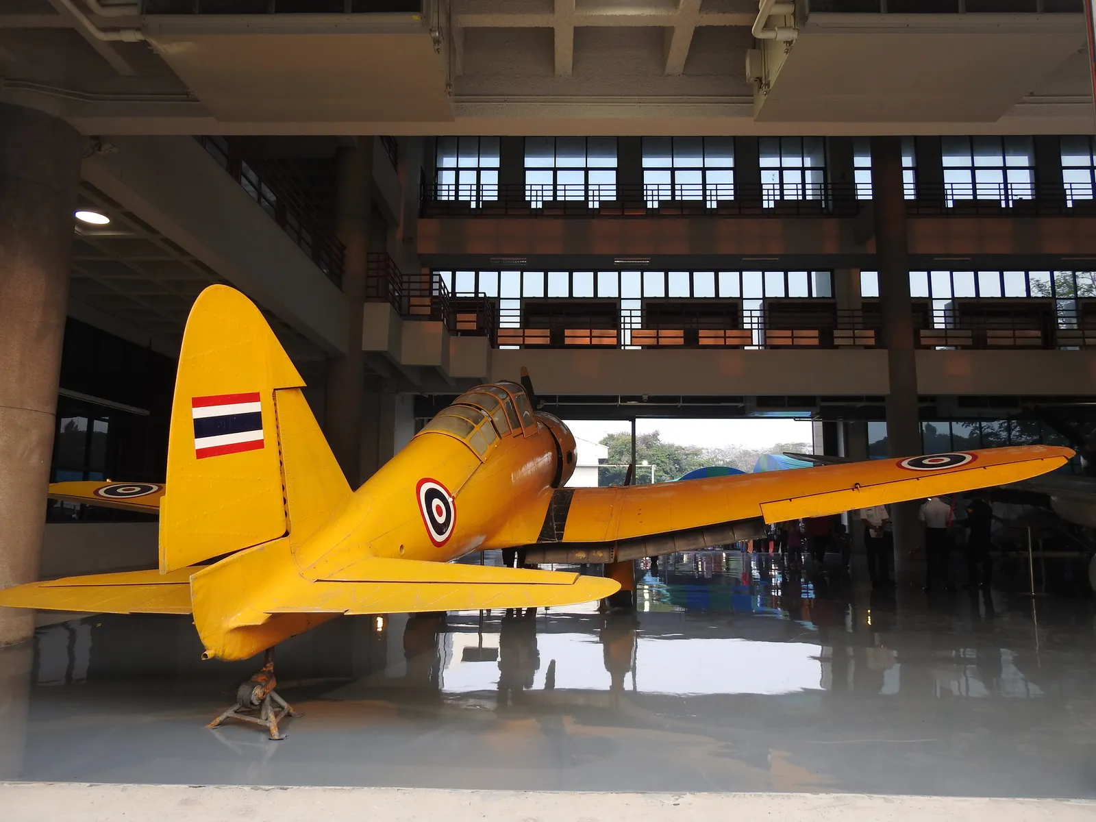 A yellow single-engine propeller aircraft with Thai roundels on its wings and tail, parked on the polished floor of a hangar at the Royal Thai Air Force Museum in Bangkok