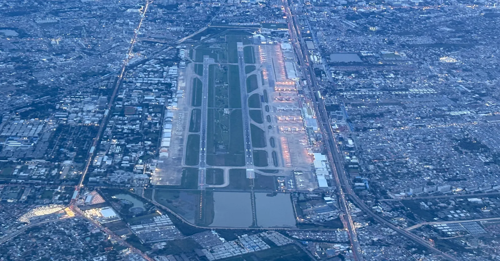 Don Mueang Airport in Bangkok seen from the air at dusk, two parallel runways with a long green strip between them and the terminals and city grid packed tight along both sides